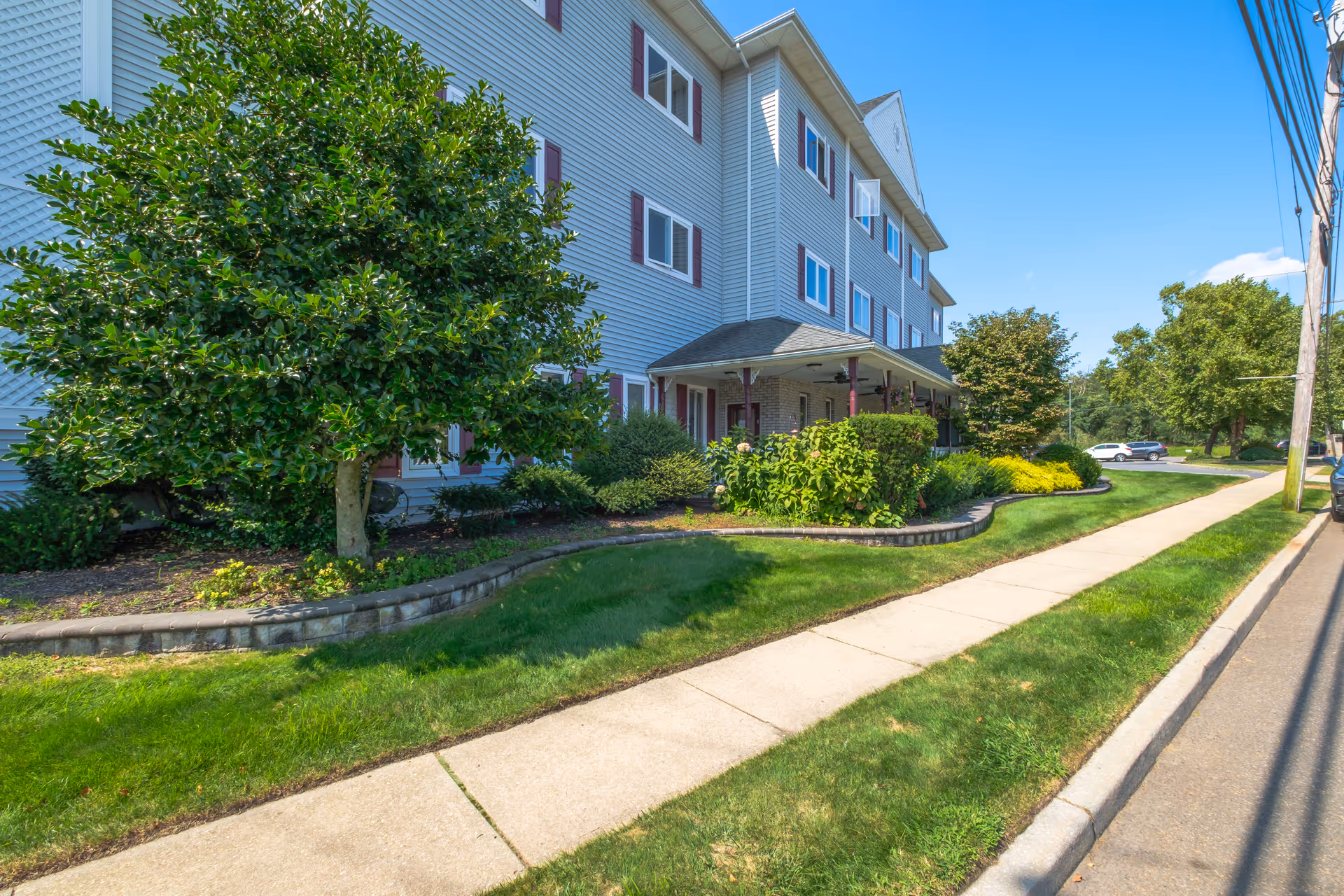 Three-story light-gray siding building with a covered entrance, landscaped front lawn, and a sidewalk running along the street.