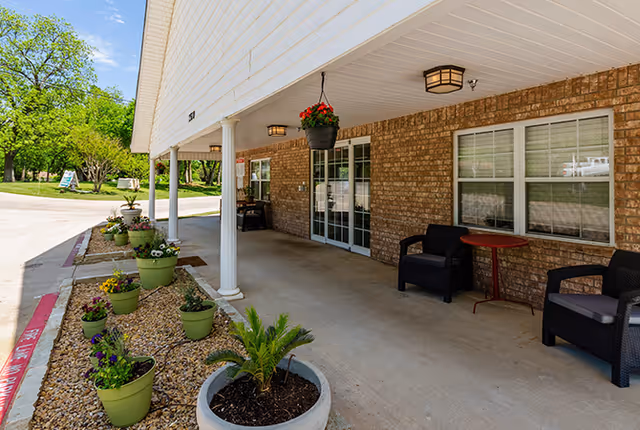Covered outdoor patio area of a brick building with white columns, hanging flower pot, several potted plants along the edge, two black chairs and a small red table near windows, and a concrete walkway leading to a green lawn and trees in the background.