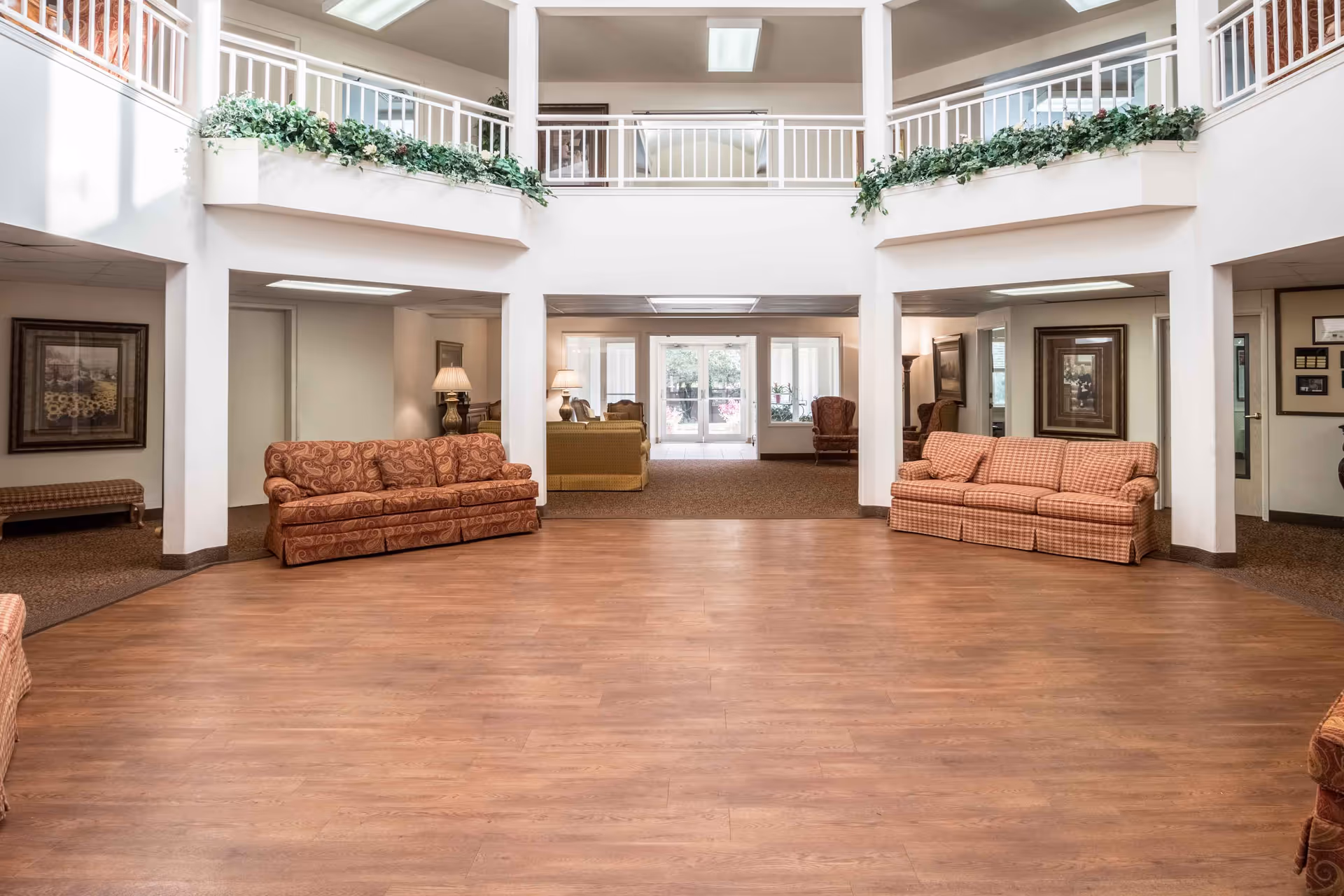 Spacious and well-lit common area in a senior living facility with wooden flooring, multiple patterned sofas, armchairs, lamps, framed artwork on the walls, and a second-floor balcony with white railings and green plants.