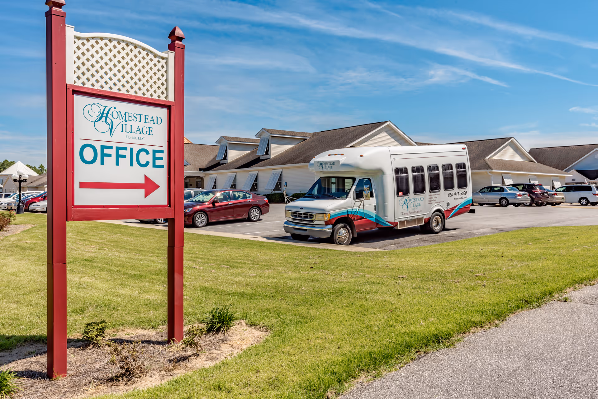 Exterior view of Homestead Village of Pensacola showing a red and white sign indicating the office direction with an arrow. In the background, there is a parking lot with several cars and a white shuttle bus with the facility's logo parked near a single-story building under a blue sky.