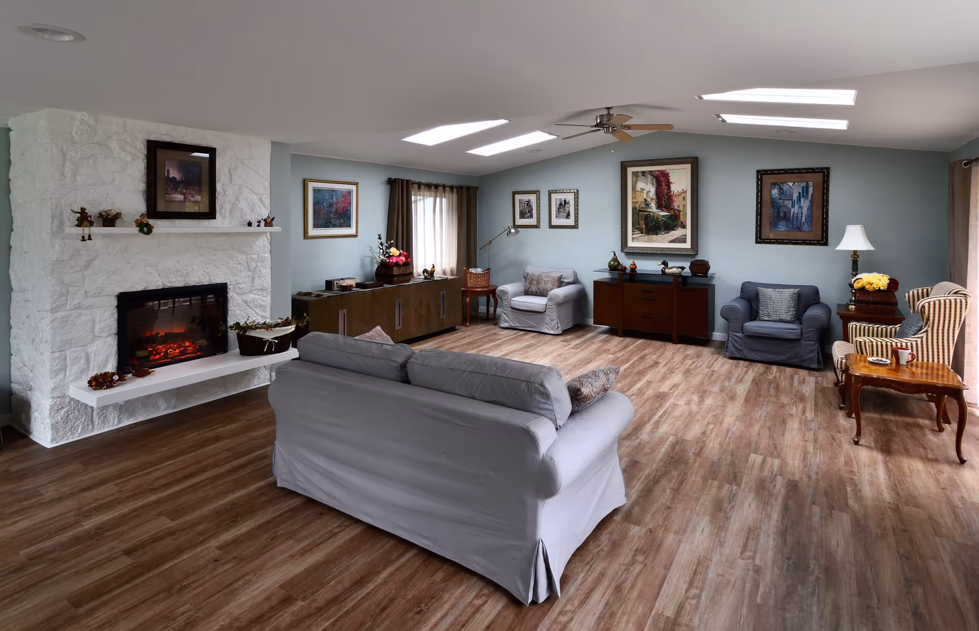 A cozy senior living room with wooden flooring, a white stone fireplace with a lit electric fire, a gray sofa, two armchairs, a striped chair, wooden side tables, and various framed paintings on light blue walls. The room is well-lit with natural light from skylights and windows with brown curtains.