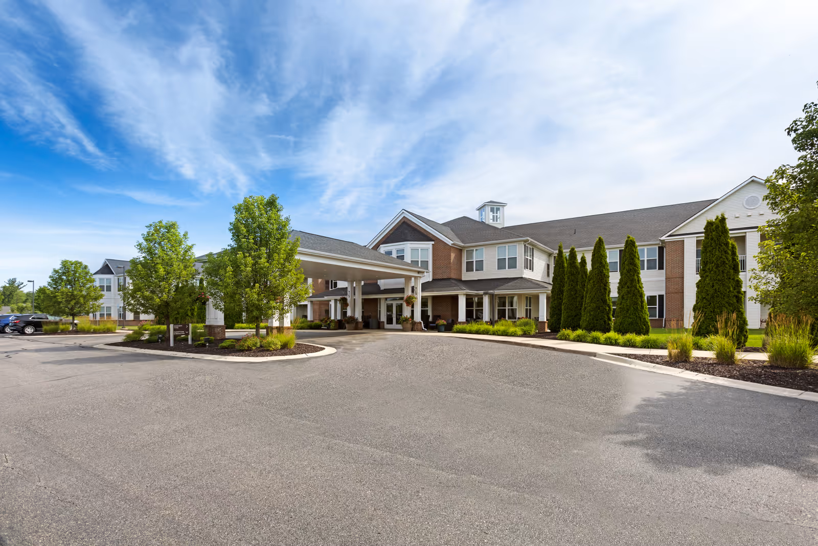 Front entrance of a large two-story senior living facility with a covered porte-cochere, landscaped beds, and a parking area under a blue sky.