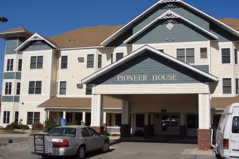 Front entrance of a multi-story senior living building with a covered porte-cochere labeled "Pioneer House" and parked cars.
