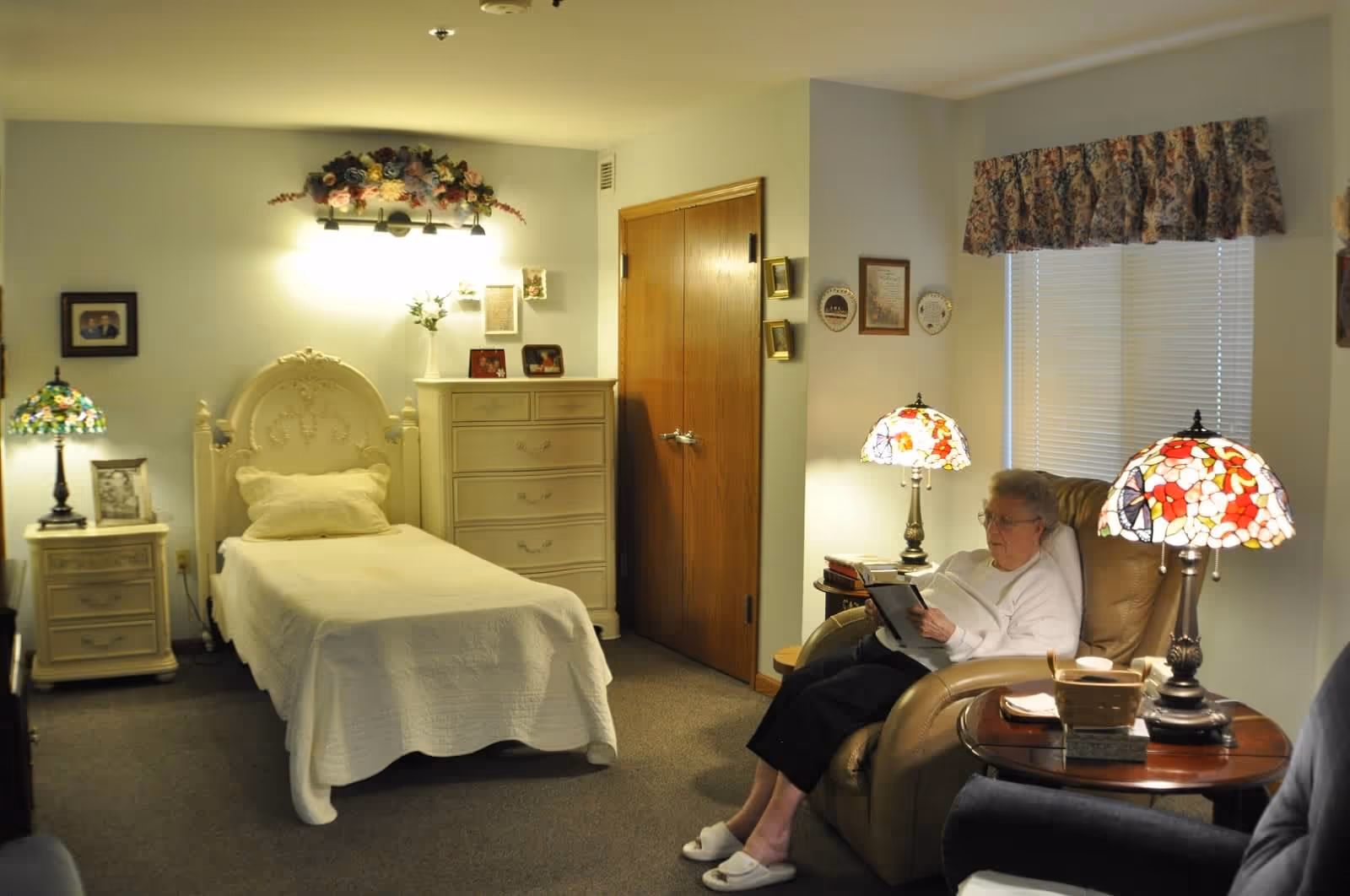 A cozy bedroom in a senior living facility with a single bed covered in a white quilt, a matching nightstand and dresser, floral wall decor above the bed, and a woman sitting in a recliner chair reading a book. The room is softly lit by two stained glass table lamps and has a window with floral valance curtains.