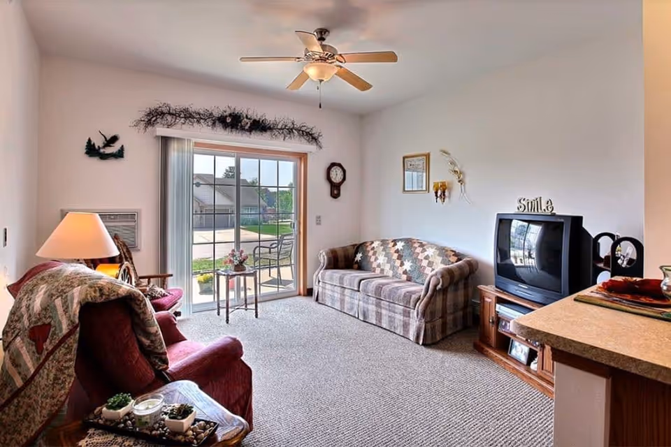 A cozy living room with a plaid sofa and a maroon armchair covered with a quilt. There is a wooden side table with a lamp and decorative items, a small TV on a wooden stand, a ceiling fan, and a sliding glass door leading to an outdoor patio with chairs. The walls have a clock, framed pictures, and decorative wall hangings.