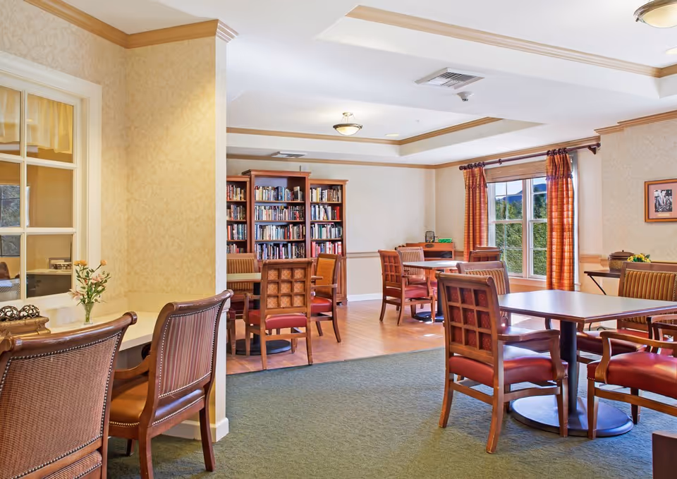 A well-lit common area in a senior living facility featuring several wooden tables and chairs with red cushions. There is a bookshelf filled with books against the back wall, a window with orange curtains letting in natural light, and a small desk area with a vase of flowers on the left side.