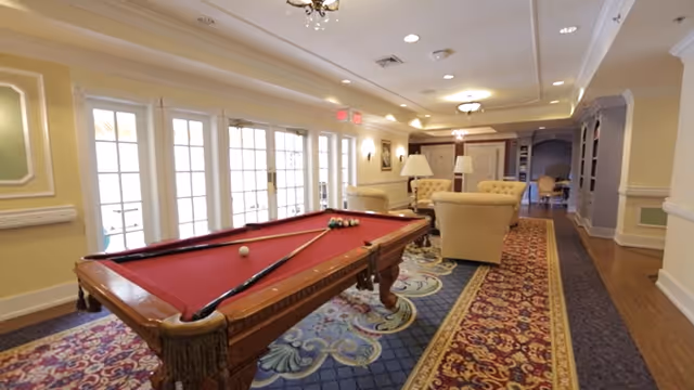 A spacious and well-lit common area featuring a red felt pool table with cues and balls arranged on it. The room has elegant patterned carpets, cream-colored armchairs, and lamps. Large windows with multiple panes allow natural light to fill the space, and the ceiling has recessed lighting and decorative molding.