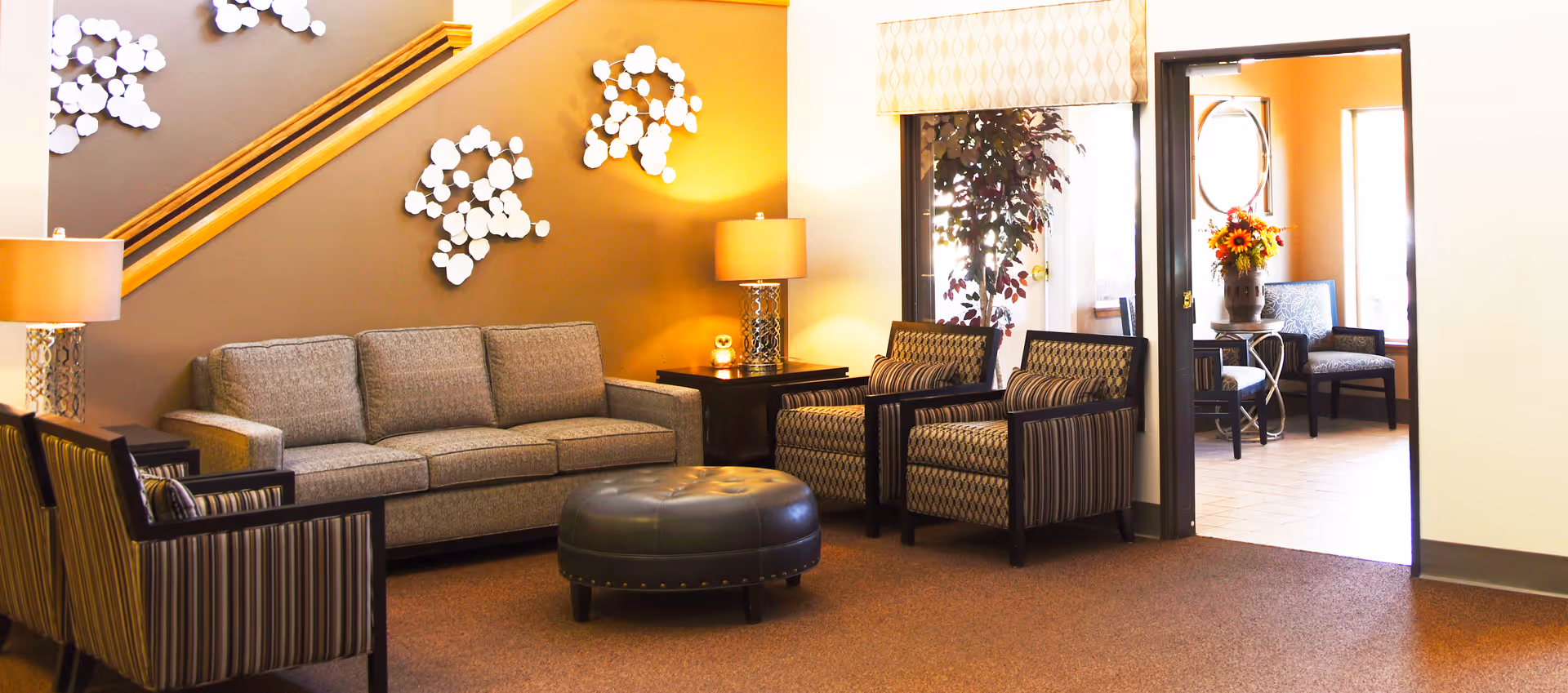 A cozy living room area with a gray sofa, two patterned armchairs, a round black ottoman, and two side tables with lamps. The wall behind the sofa is decorated with white circular wall art and a wooden handrail. There is an open doorway leading to another room with chairs and a vase of flowers on a table.