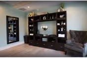 A cozy interior room featuring a large dark wood entertainment center with shelves displaying decorative items and a vase with flowers in the center. To the right of the entertainment center is a plush gray armchair, and to the left is a tall mirror mounted on a light green wall. The floor is light wood.