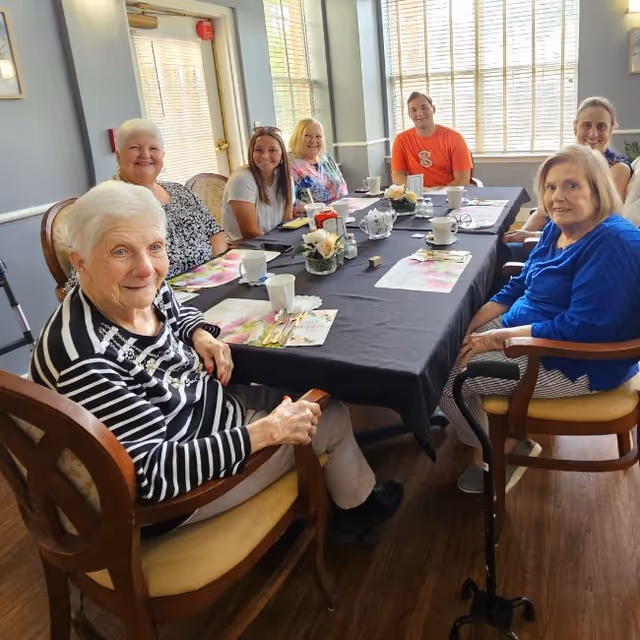 A group of older adults and caregivers sit around a table set for a meal in a bright dining room.