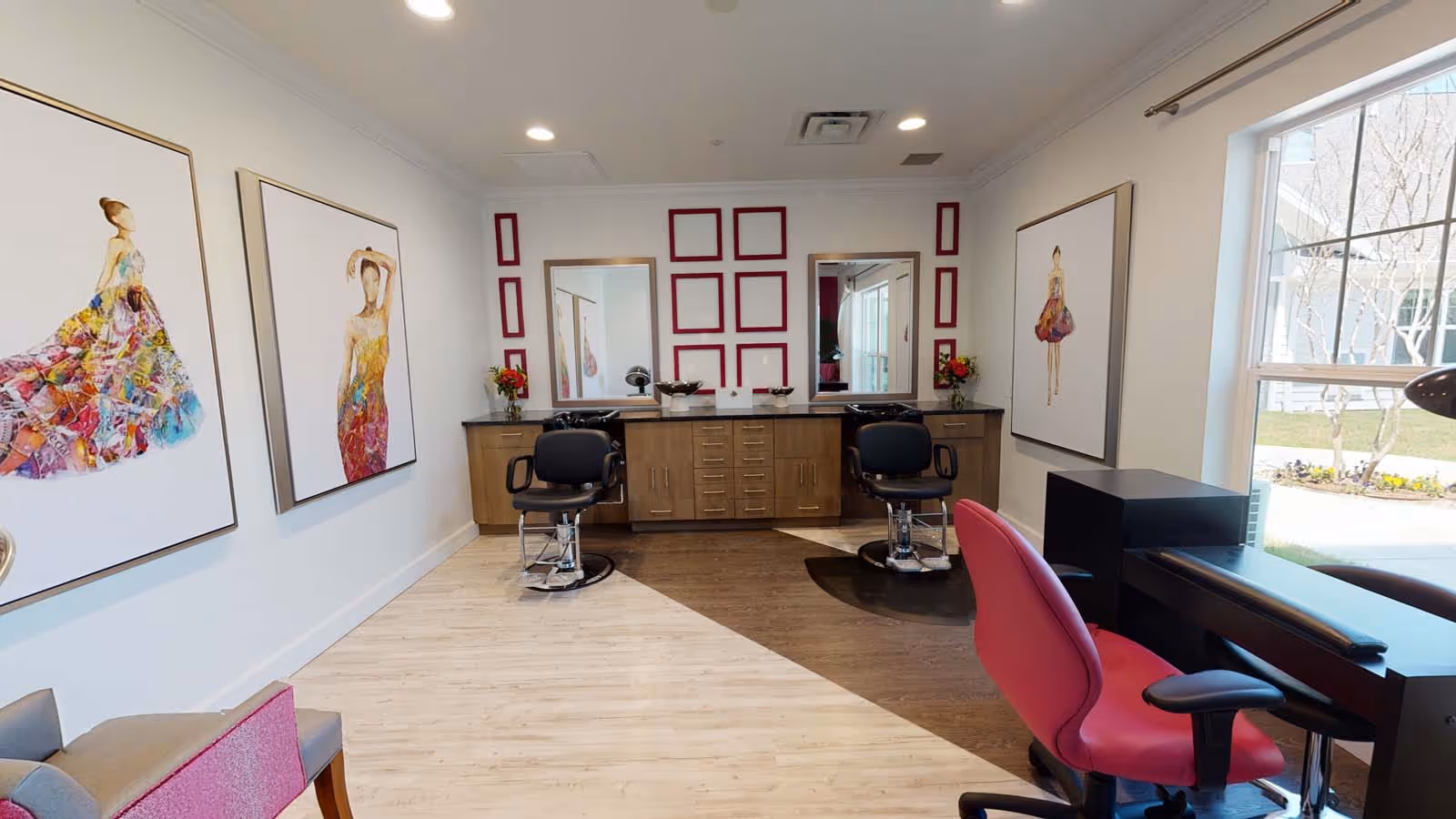 Interior view of a salon area with two black salon chairs in front of mirrors mounted on a wooden cabinet. The room has light wood flooring transitioning to darker wood near the salon chairs. There are three large framed fashion illustrations on the walls and a large window letting in natural light. A pink chair and a black manicure table are positioned near the window.
