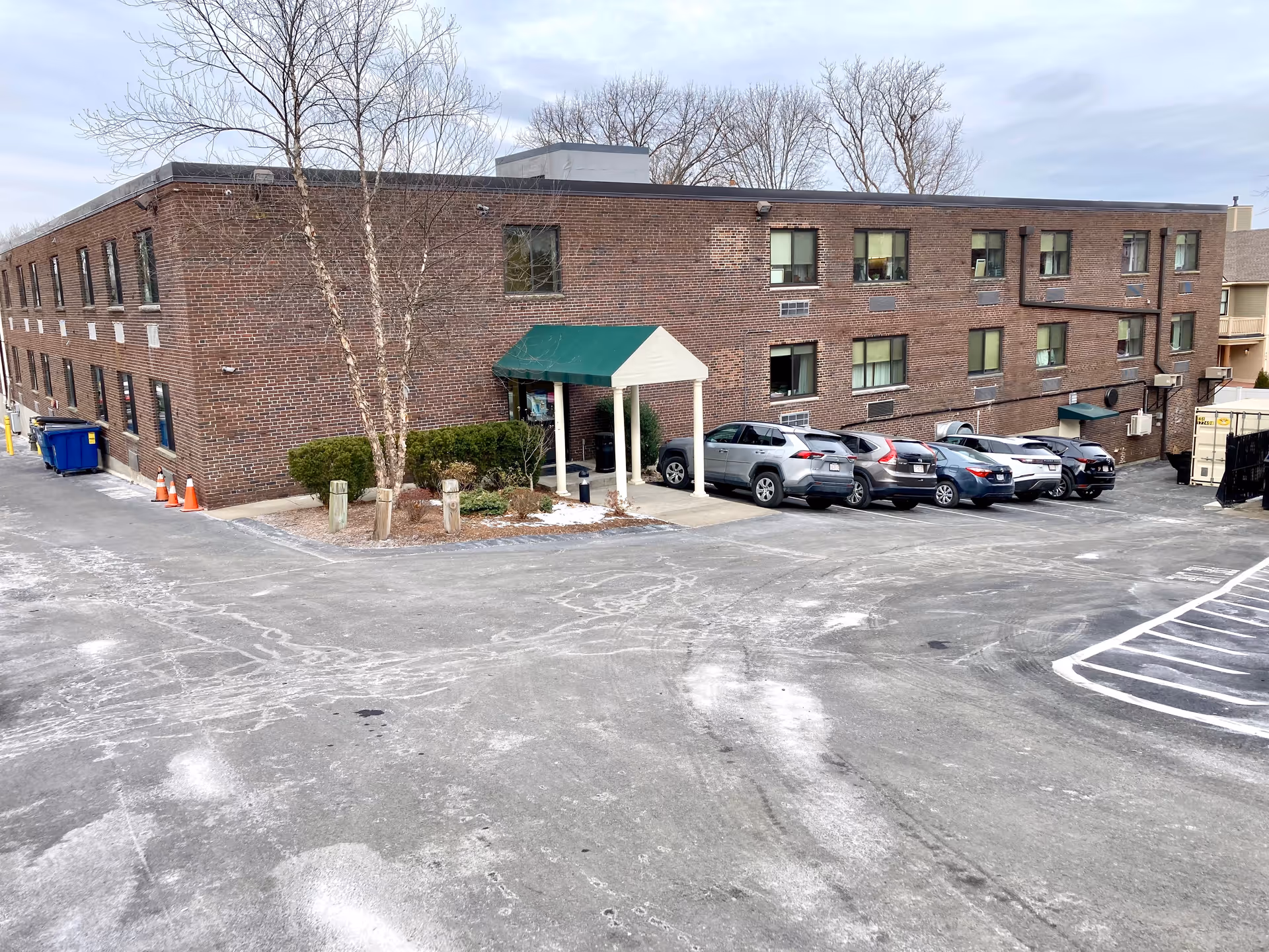 Brick two-story senior care building with a green awning entrance and parked cars in a paved parking lot.