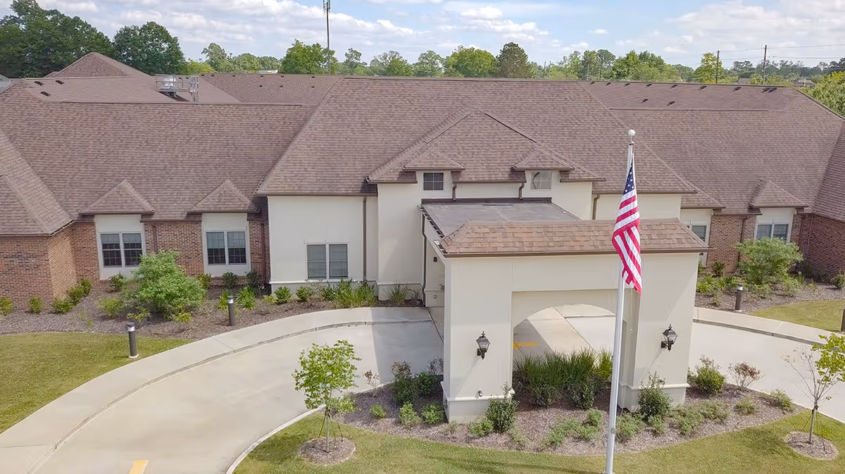 Front entrance of a senior living building with a covered porte-cochere, circular driveway, landscaping, and an American flag.