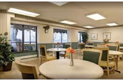 A well-lit dining room with several round tables and green cushioned chairs. Each table has a small vase with flowers. Large windows with vertical blinds allow natural light to enter. The walls are decorated with framed artwork and there are potted plants in the corners.