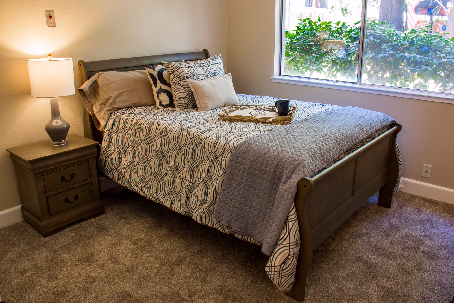 A cozy bedroom with a wooden bed frame and a patterned bedspread. There are three decorative pillows on the bed and a tray with a cup and a book. A wooden nightstand with two drawers and a lamp is beside the bed. A large window shows greenery outside, letting in natural light.