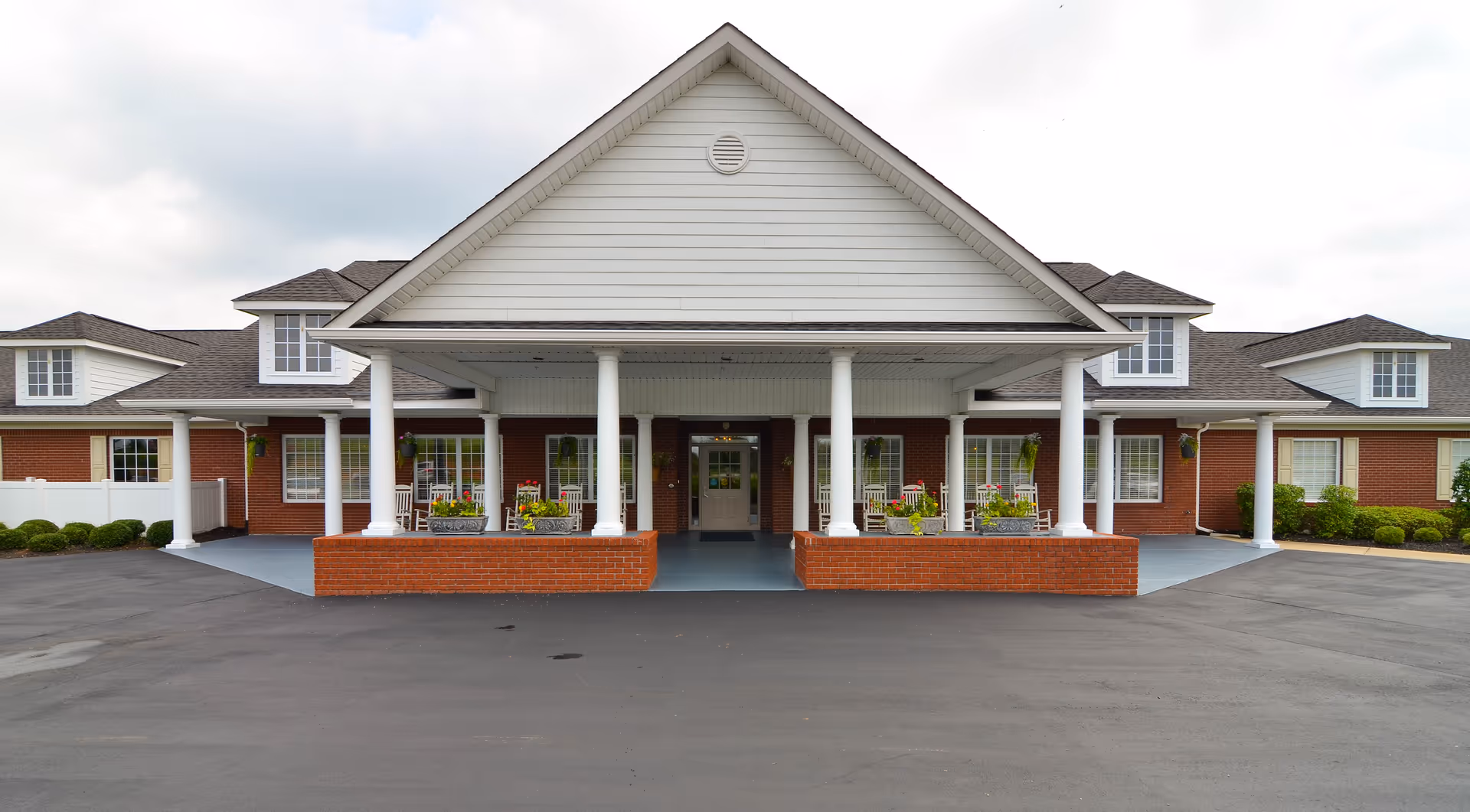 Front exterior view of a single-story senior living facility building with a large covered entrance supported by white columns, brick walls, multiple windows, and a paved driveway in front.