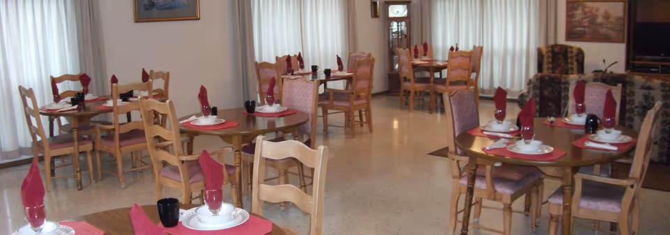 Dining room with several round wooden tables set with plates, black cups and folded red napkins and upholstered chairs.