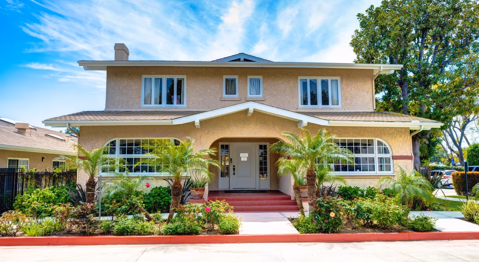 Front exterior view of a two-story beige building with large windows, a covered entrance with steps, and landscaped greenery including palm trees and bushes in front under a blue sky with some clouds.