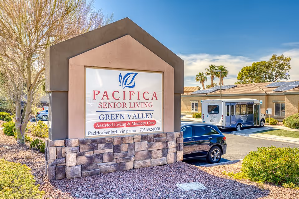 Outdoor view of the entrance sign for Pacifica Senior Living Green Valley, an assisted living and memory care facility. The sign is mounted on a stone and stucco base with landscaping around it. In the background, there is a parking lot with a black car and a shuttle bus, along with a building with solar panels on the roof and palm trees under a clear blue sky.