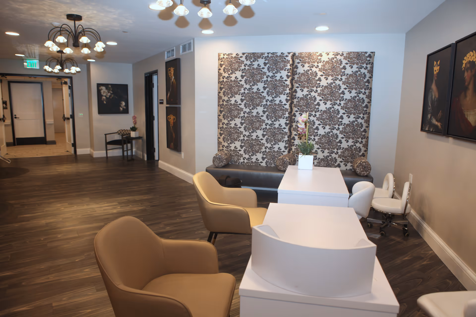 Interior view of a senior living facility lobby or common area with modern beige armchairs, white desks, floral patterned wall panels, decorative paintings on the walls, and overhead lighting fixtures. The floor is dark wood, and there is a hallway with doors and an exit sign in the background.