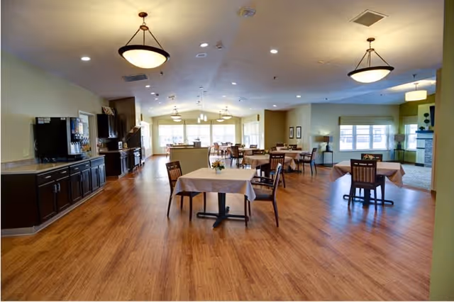 Spacious dining room with wooden flooring, several tables covered with light-colored tablecloths, and wooden chairs. The room is well-lit with ceiling lights and natural light from large windows. There is a kitchen area with dark cabinets and appliances on the left side.