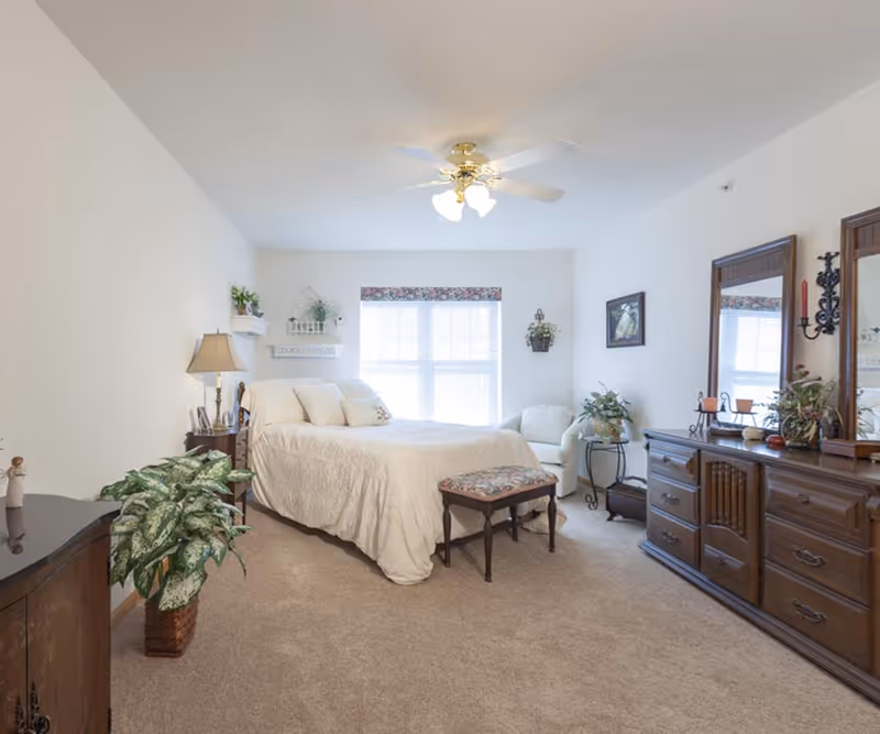A bright and cozy bedroom with a large window covered by blinds and a floral valance. The room features a bed with white bedding and pillows, a wooden nightstand with a lamp, a small upholstered bench at the foot of the bed, a white armchair, and a wooden dresser with two mirrors. There are several plants and decorative items placed around the room, and a ceiling fan with lights is mounted on the ceiling.