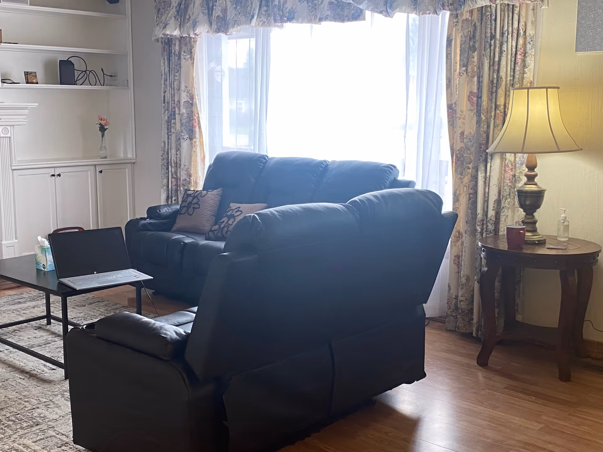 A cozy living room with two dark leather sofas, a coffee table with a laptop and tissue box, a side table with a lamp, a cup, and a bottle of hand sanitizer. The room has floral curtains and a built-in white cabinet with shelves and a small vase with a flower.