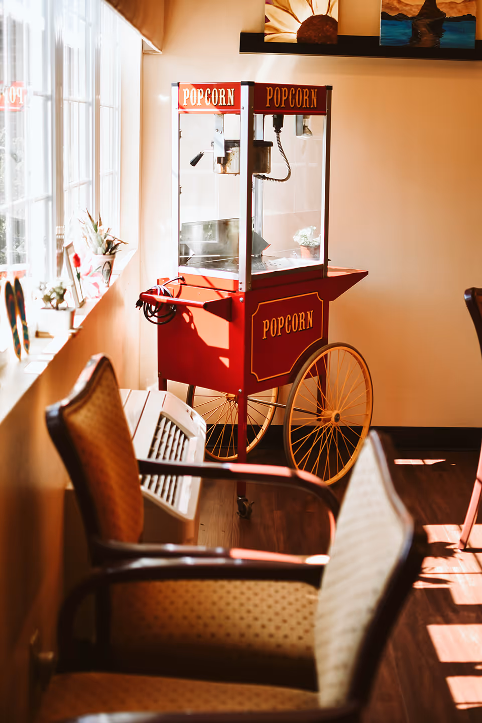 A red popcorn cart on wheels sits in a sunlit interior seating area by windows with chairs.