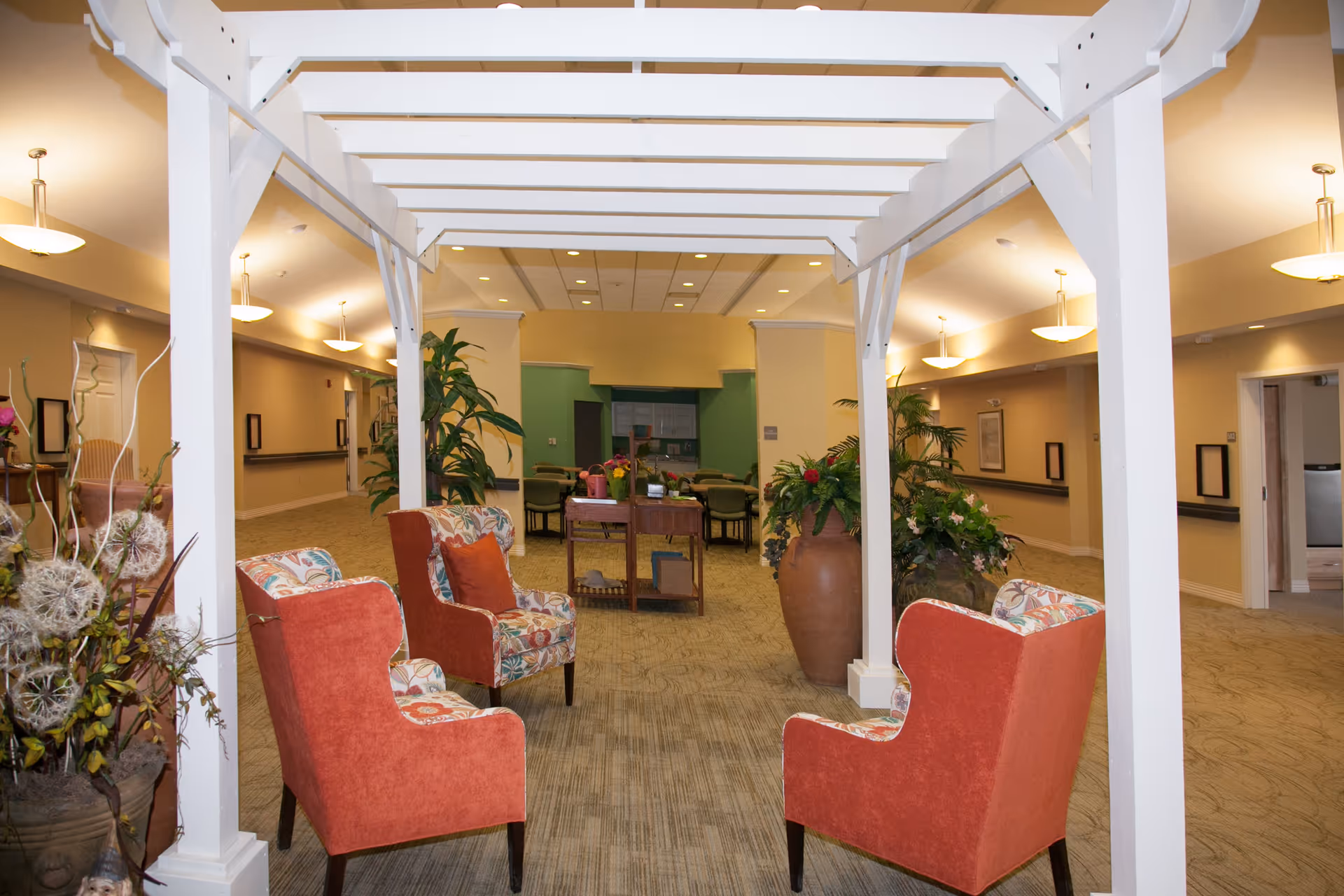 A cozy indoor seating area in a senior living facility with four red upholstered armchairs arranged under a white wooden pergola structure. The space is decorated with large potted plants and floral arrangements, with a table and additional chairs visible in the background. The walls are painted in warm tones and the area is well-lit with ceiling lights.