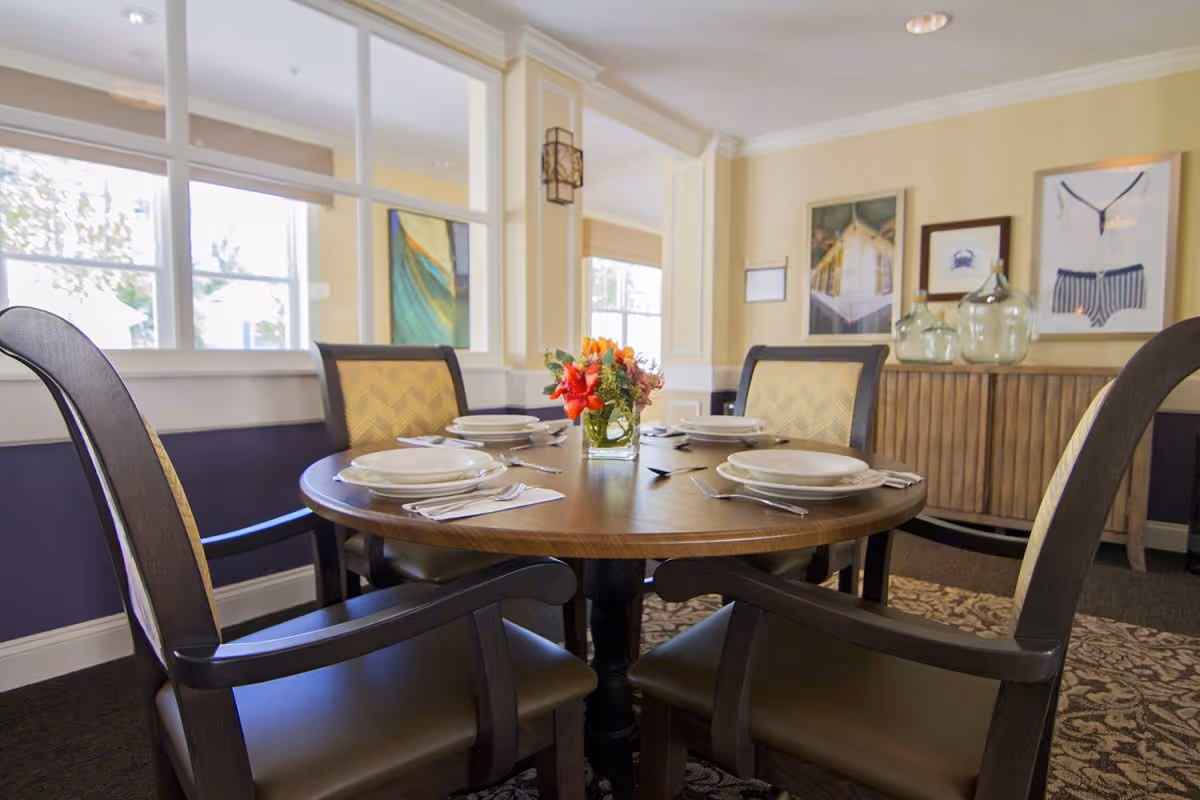 A round wooden dining table set for four with plates, napkins, and silverware, featuring a small floral centerpiece. The room has beige walls with framed artwork and large windows letting in natural light. Four cushioned chairs with wooden frames surround the table.