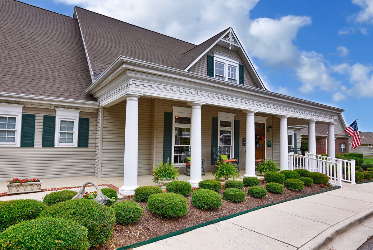 Exterior view of Columbia Cottage Florence, showing a beige building with white columns supporting a covered porch. The porch has rocking chairs and potted plants. There are neatly trimmed bushes and a small garden in front, with an American flag displayed near the entrance under a partly cloudy sky.