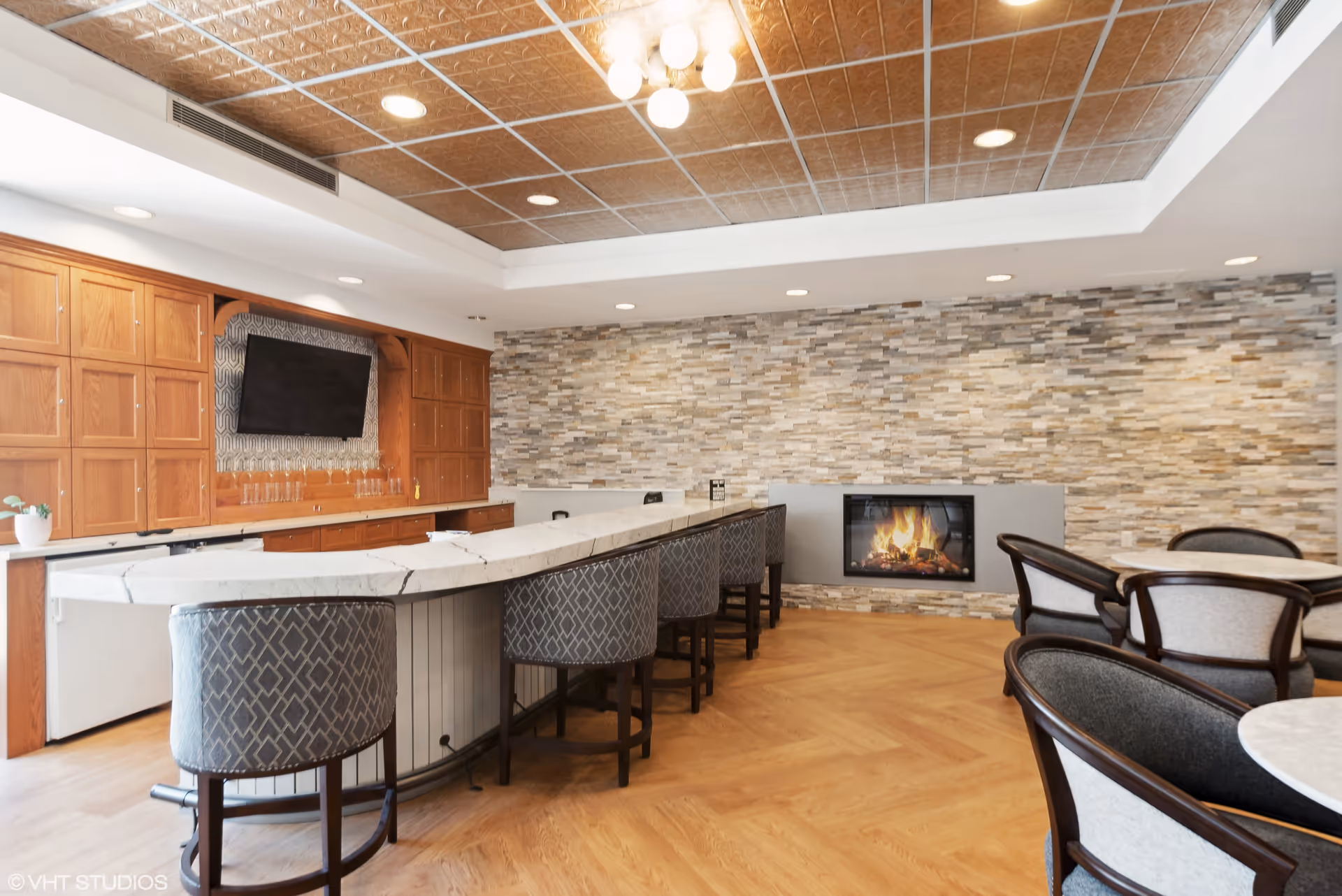 Interior view of a lounge area with a long marble countertop bar and several upholstered bar stools. The back wall features wooden cabinets and a mounted flat-screen TV. There is a modern fireplace set in a stone accent wall, and round tables with cushioned chairs are arranged nearby. The ceiling has a decorative tile pattern with recessed lighting and a central light fixture.