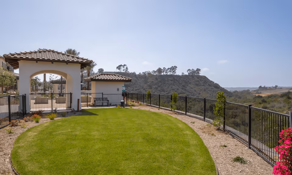 Outdoor area at Santianna Oakmont Signature Senior Living featuring a well-maintained green lawn, black metal fencing, a small pavilion with a tiled roof, benches, and scenic views of hills and trees under a clear blue sky.
