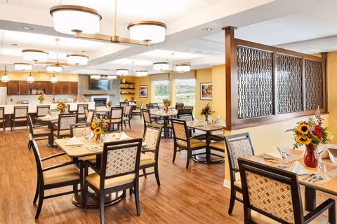 A bright and spacious dining room with multiple wooden tables and chairs arranged neatly. Each table is set with napkins, glasses, and small floral centerpieces. The room features warm lighting from ceiling fixtures and pendant lights above a counter area. The walls are decorated with framed artwork and a decorative wooden partition separates part of the space. Large windows allow natural light to fill the room.