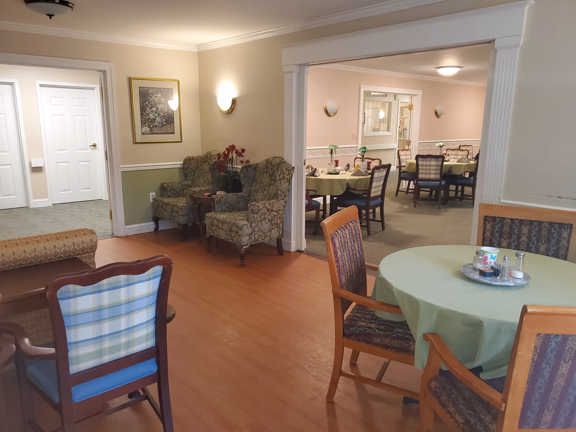 A cozy communal interior showing round dining tables with green tablecloths, wooden chairs, and upholstered armchairs beside a connected dining room.
