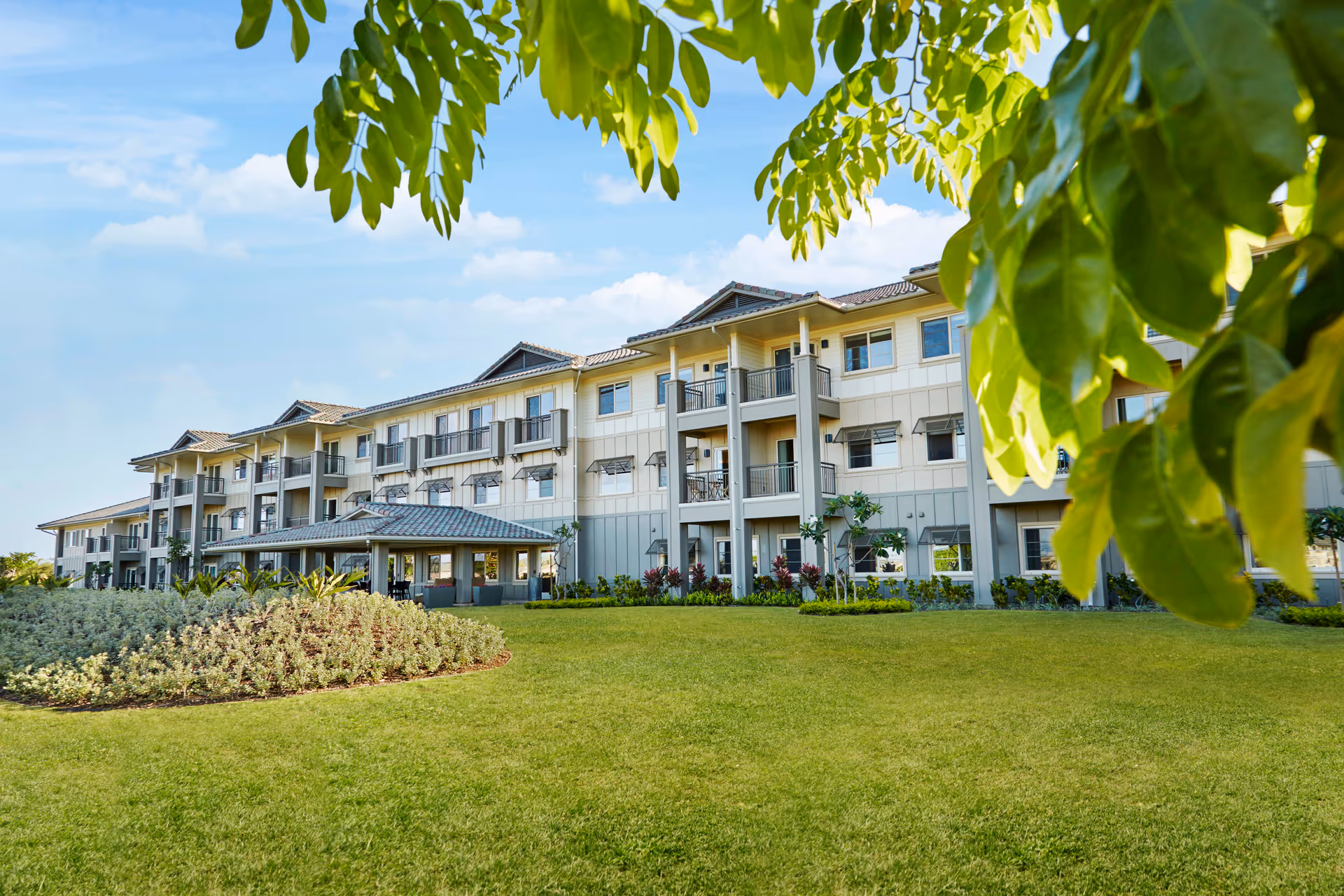 Exterior view of a three-story senior living facility building with balconies, surrounded by a well-maintained lawn and landscaping, with tree leaves partially framing the top of the image under a blue sky with some clouds.