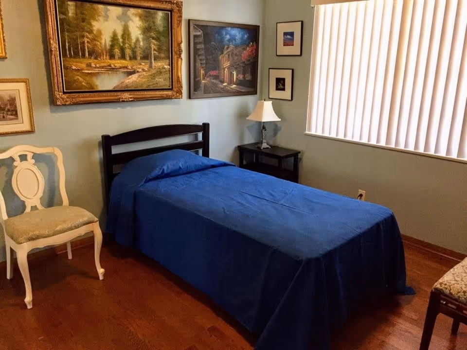 A small bedroom with a single bed covered in a blue bedspread, a white decorative chair, a dark wooden nightstand with a lamp, and several framed paintings on light blue walls. A window with vertical blinds lets in natural light.