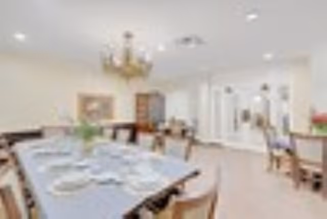 A bright dining room with multiple wooden tables and chairs arranged neatly. The tables are set with white plates and napkins, and a chandelier hangs from the ceiling. The room has light-colored walls and flooring, with a cabinet and framed artwork visible in the background.