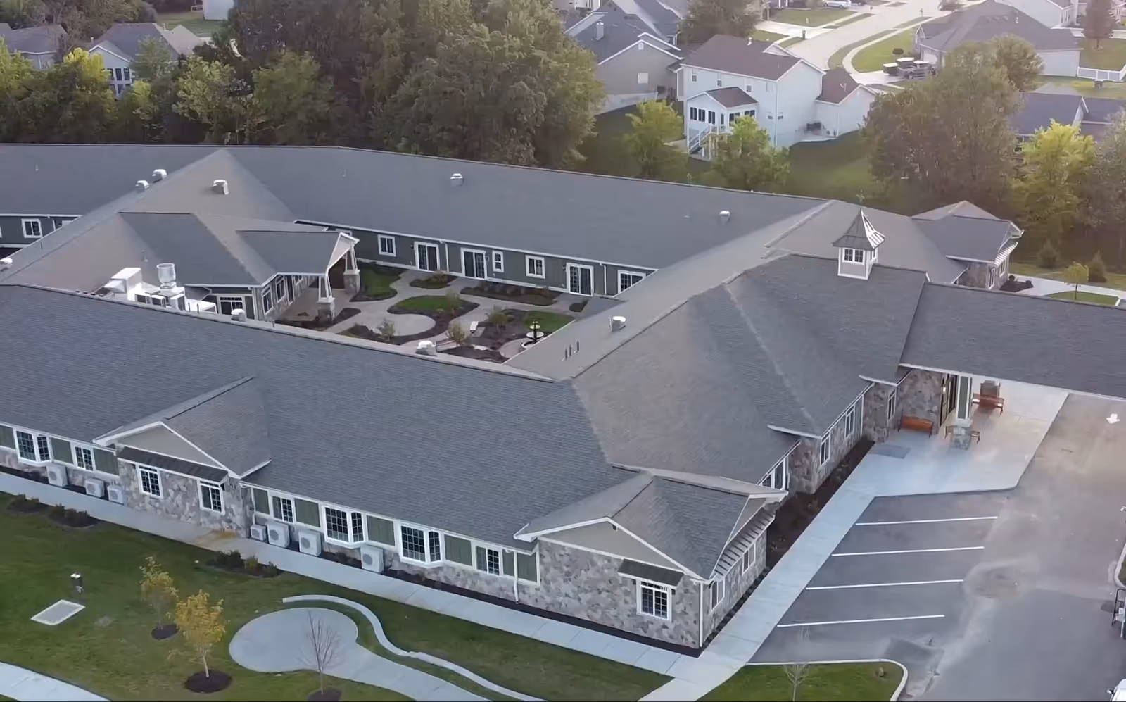 Aerial view of a single-story assisted living facility with a large, gray shingled roof and stone exterior walls. The building is arranged in a U-shape around a landscaped courtyard with pathways and greenery. There is a parking area adjacent to the building, and residential houses and trees are visible in the background.