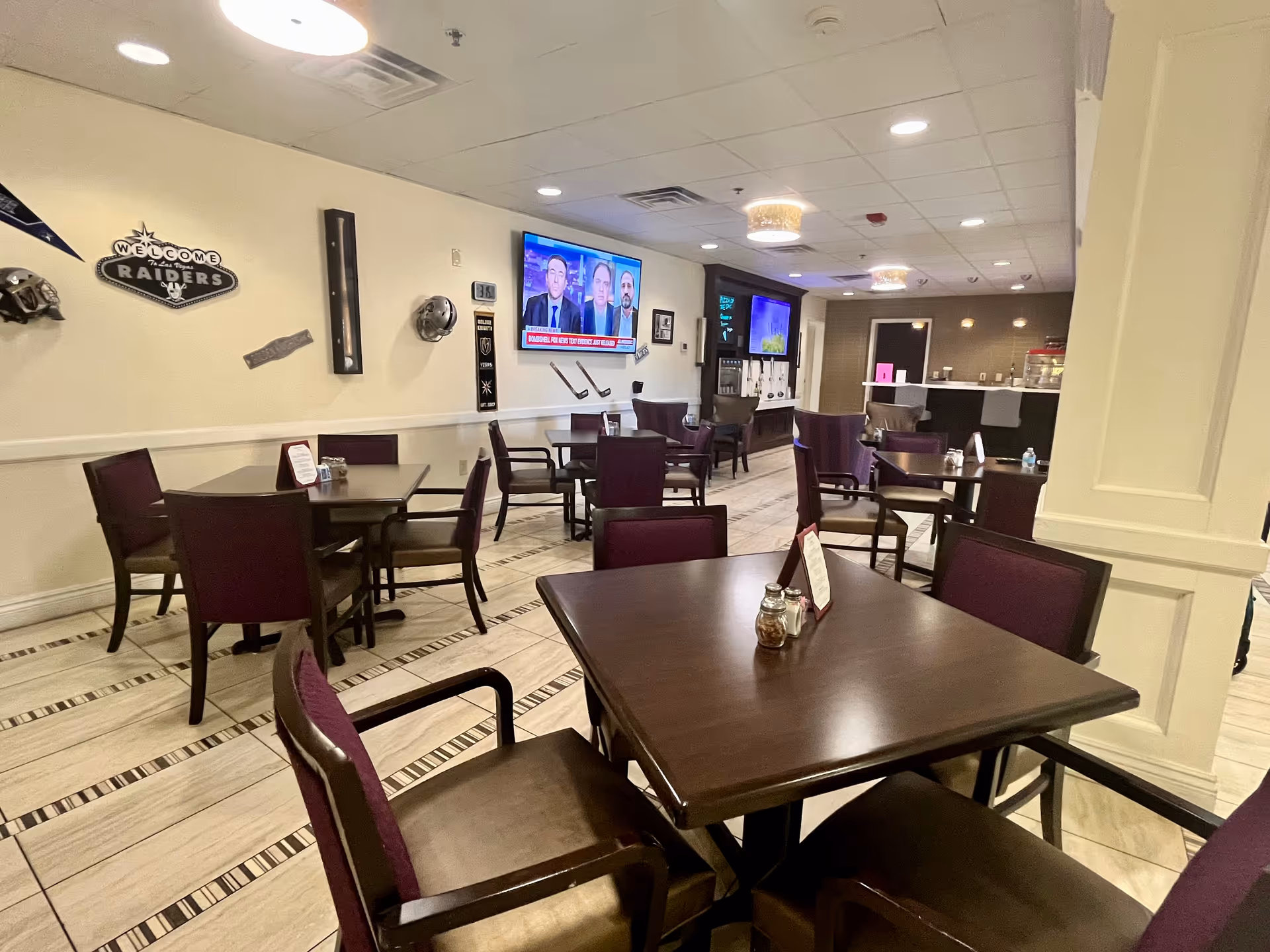 Interior view of a dining area with several dark wooden tables and chairs with purple cushions. The walls are decorated with sports memorabilia including a 'Welcome Raiders' sign and helmets. A flat-screen TV is mounted on the wall showing a news program. In the background, there is a beverage station with dispensers and a counter.