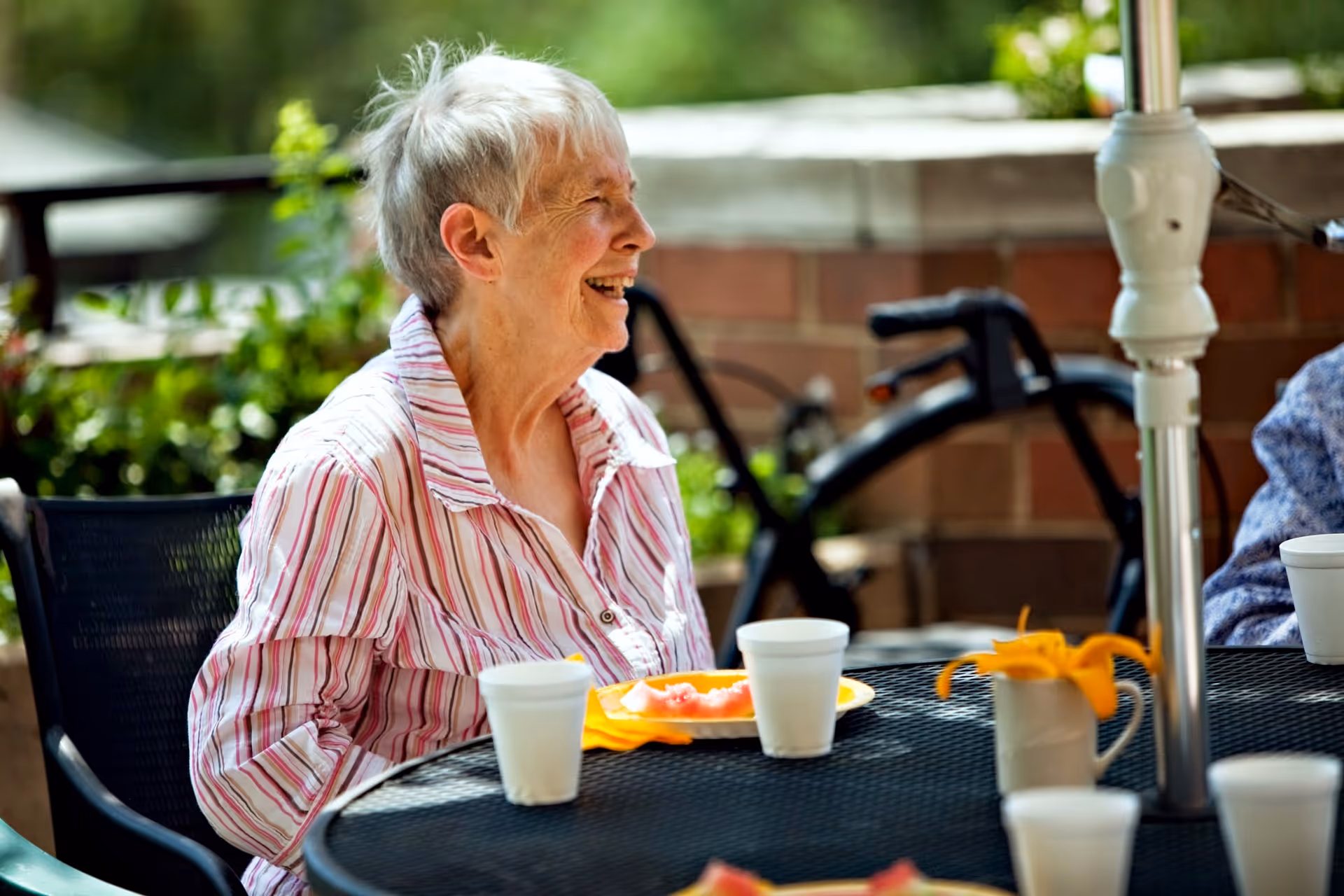 An elderly woman with short white hair wearing a striped shirt is sitting at a round outdoor table with a black mesh surface. She is smiling and appears to be enjoying a meal or snack. On the table are white disposable cups, a plate with food, and a small white vase with orange flowers. In the background, there is a brick wall and some greenery.
