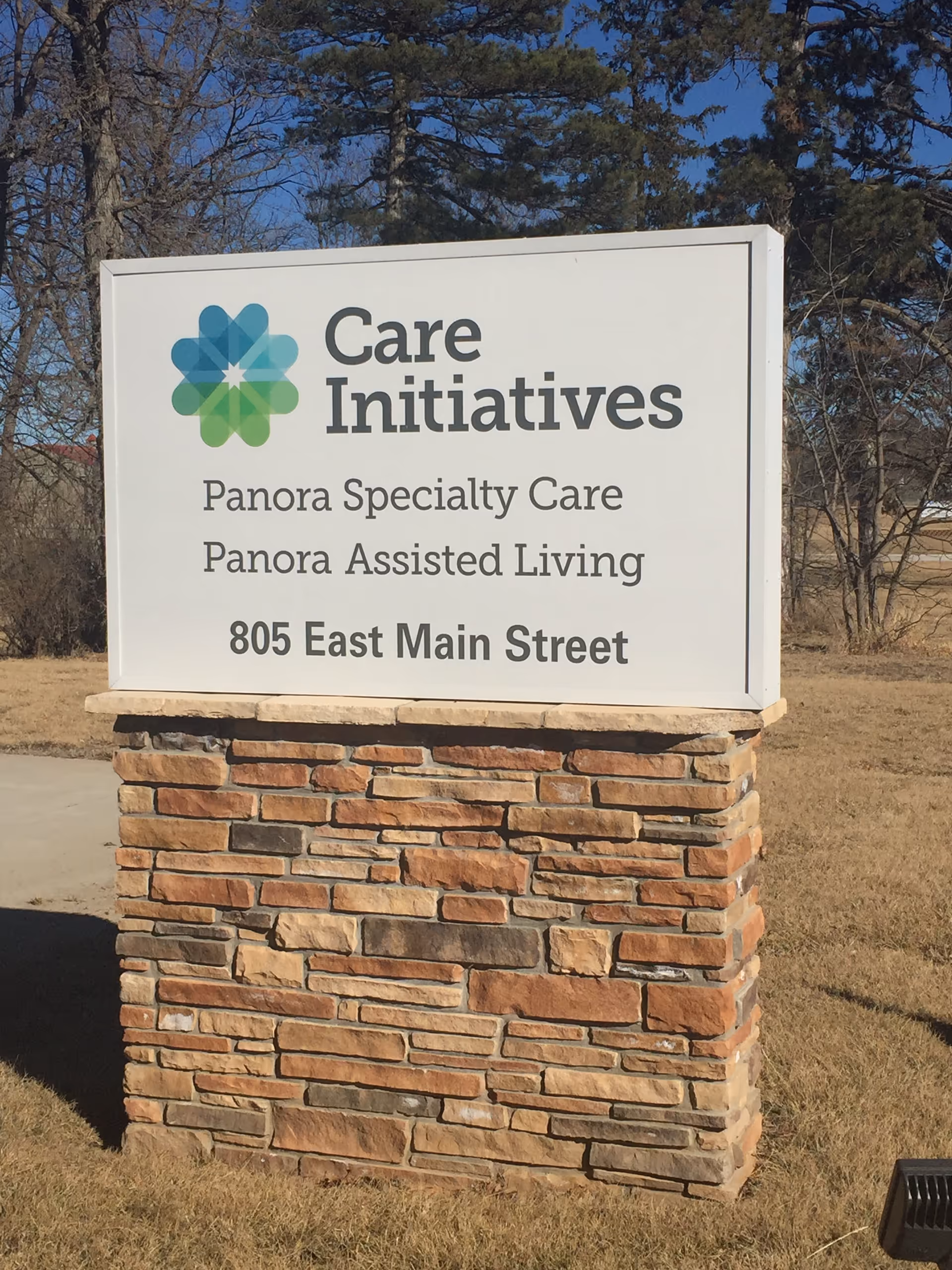 Freestanding outdoor sign on a stone base reading "Care Initiatives, Panora Specialty Care, Panora Assisted Living, 805 East Main Street" with grass and trees in the background.