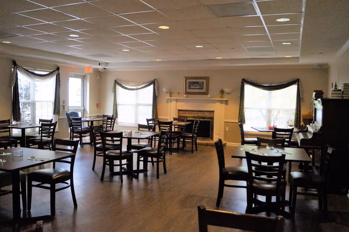 A dining room with several dark wooden tables and chairs arranged neatly. The room has large windows with curtains, a fireplace with a framed picture above it, and a piano on the right side with books stacked on top. The floor is wooden, and the ceiling has recessed lighting.