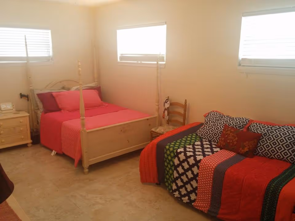 A cozy bedroom with a cream-colored four-poster bed dressed in bright pink bedding and pillows. Next to the bed is a small cream-colored nightstand with a clock and lamp. Across from the bed is a colorful sofa covered with a patchwork quilt in red, green, black, and white patterns, along with several decorative pillows. The room has beige tiled flooring and three windows with white blinds letting in natural light.