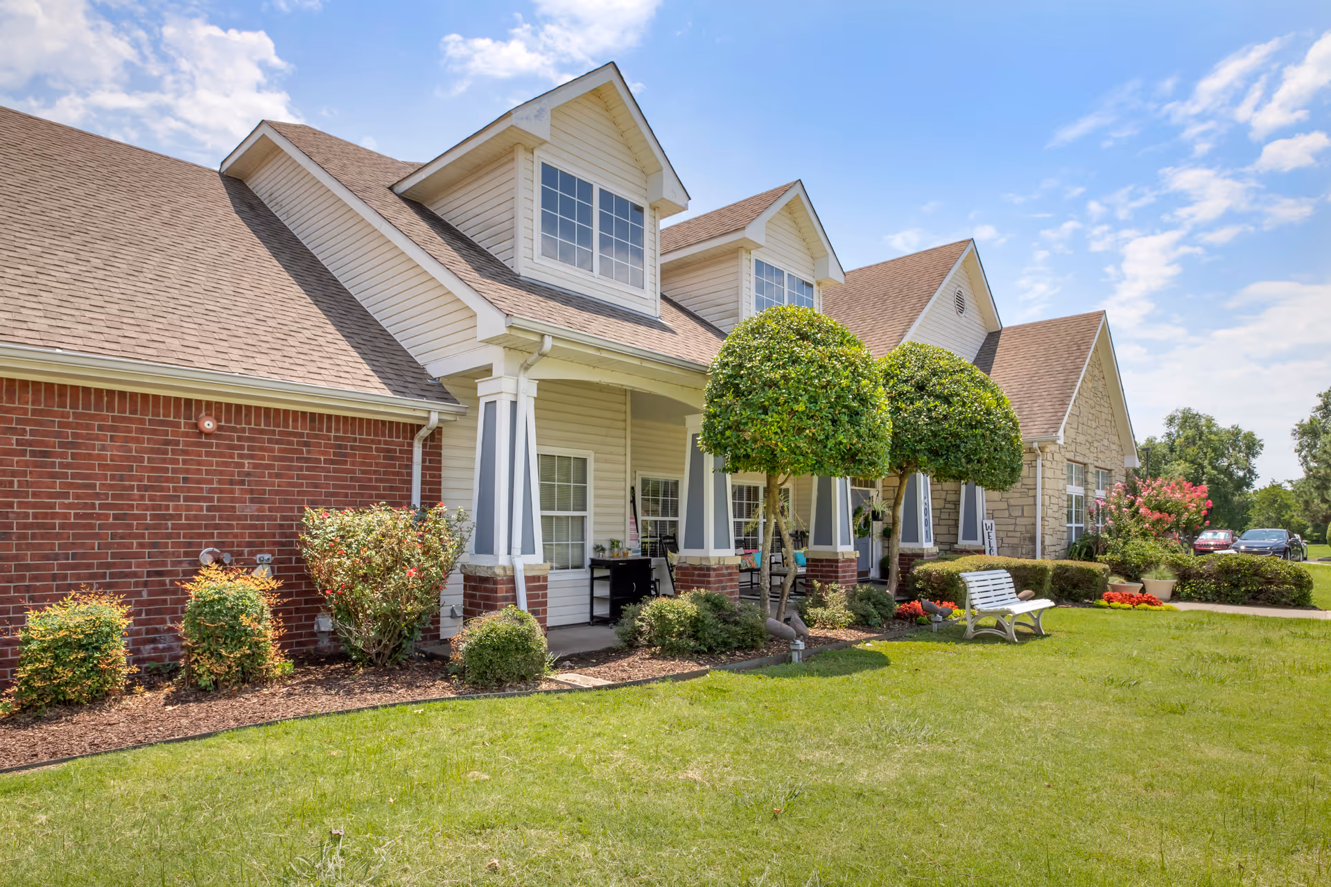 Front exterior of a senior living facility with a covered porch, brick and siding facade, trimmed trees, benches and a manicured lawn.