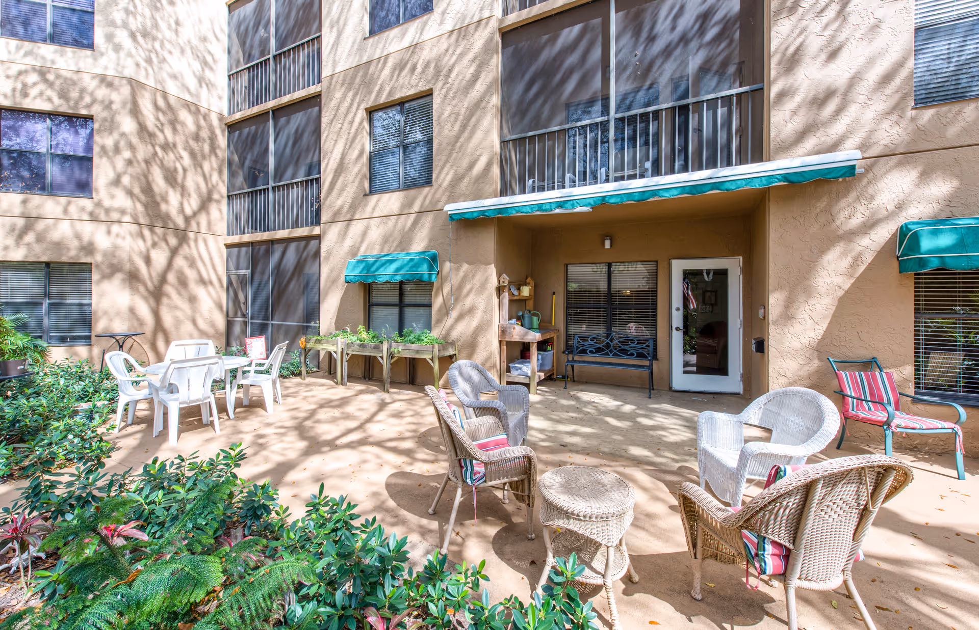 Outdoor ground-level patio courtyard with wicker and plastic chairs, tables, potted plants and green awnings in front of a multi-story building.