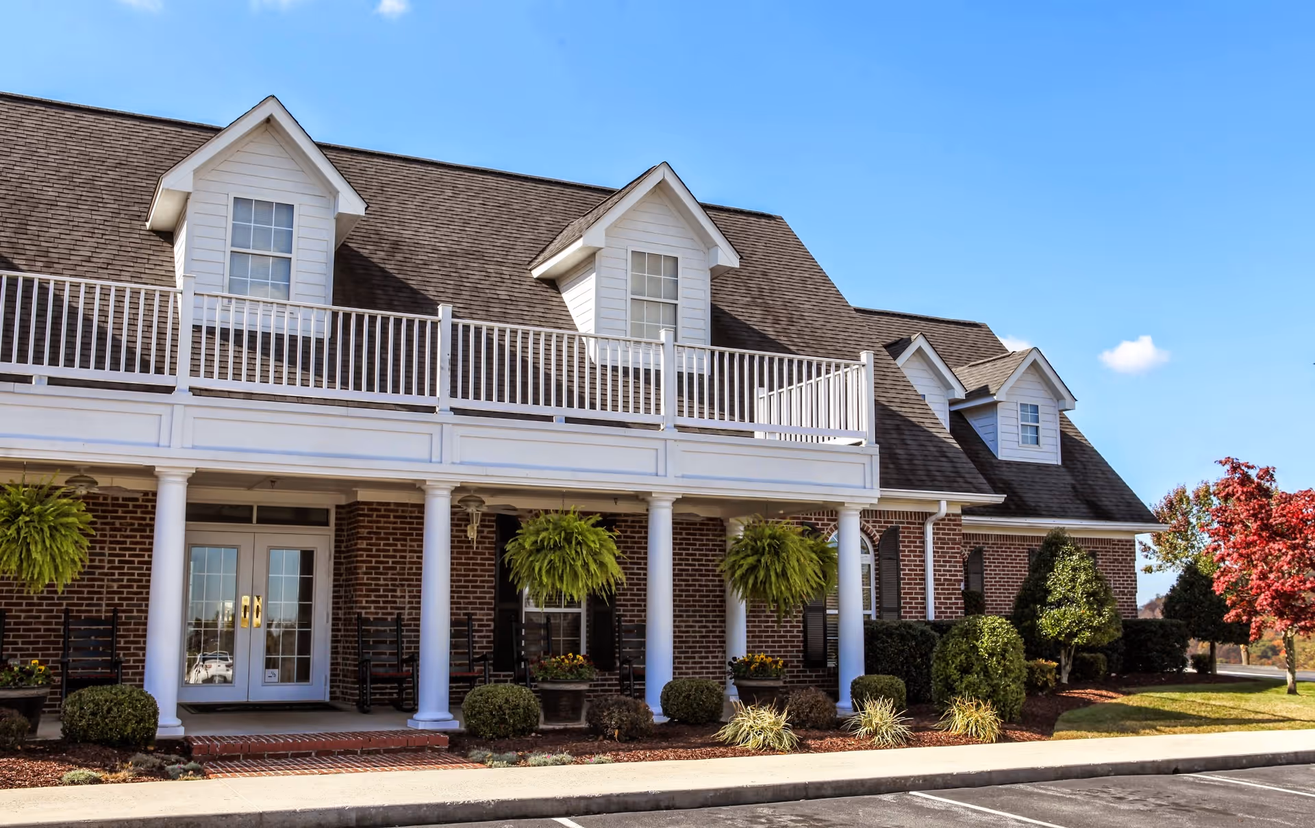 Brick two-story building with white columns, a second-story balcony, dormer windows, and landscaped entrance.