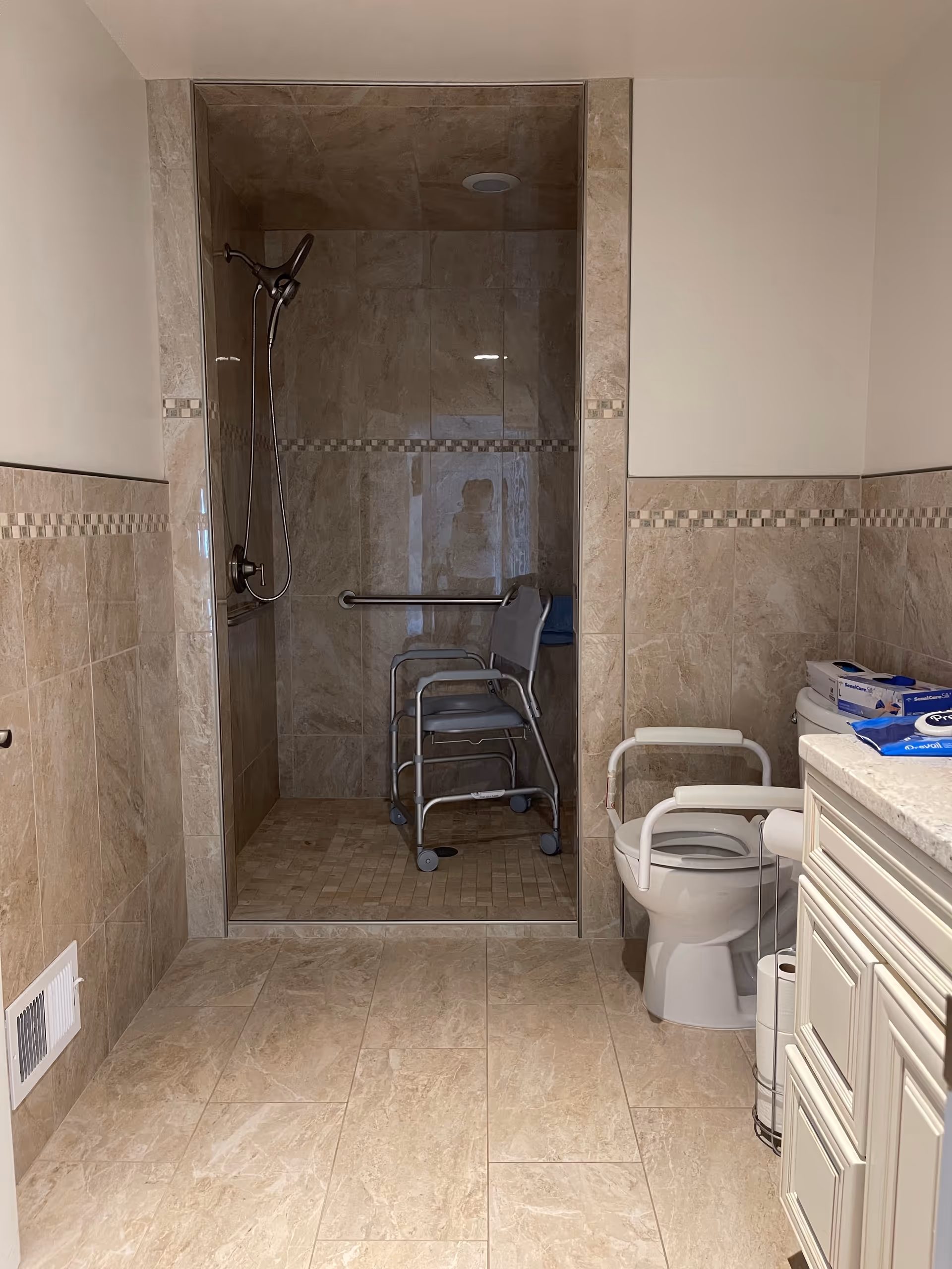 A bathroom with beige tiled walls and floor, featuring a walk-in shower with a handheld showerhead and a shower chair inside. There is a toilet with safety rails next to a white cabinet countertop, and various hygiene products are placed on the counter.