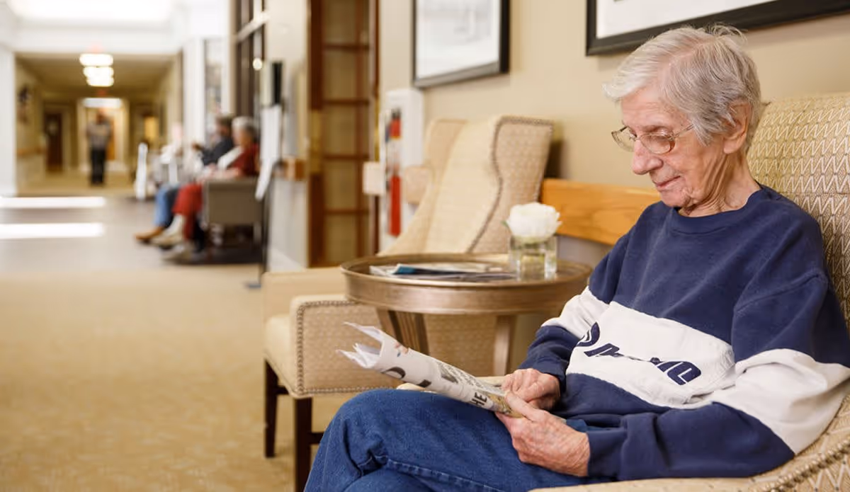 An elderly woman with short gray hair and glasses is sitting on a cushioned bench in a hallway, reading a newspaper. Behind her is a round wooden table with a small vase containing a white flower. The hallway has several chairs along the wall, and other elderly individuals are seated further down the corridor.