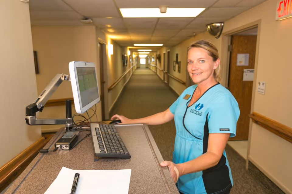 A smiling female healthcare worker in a blue uniform stands at a computer workstation in a well-lit hallway of a healthcare facility. The hallway has handrails on both sides and several doors along the walls. The worker is looking at the camera, and the uniform has the logo and name 'Beatrice Health and Rehabilitation'.